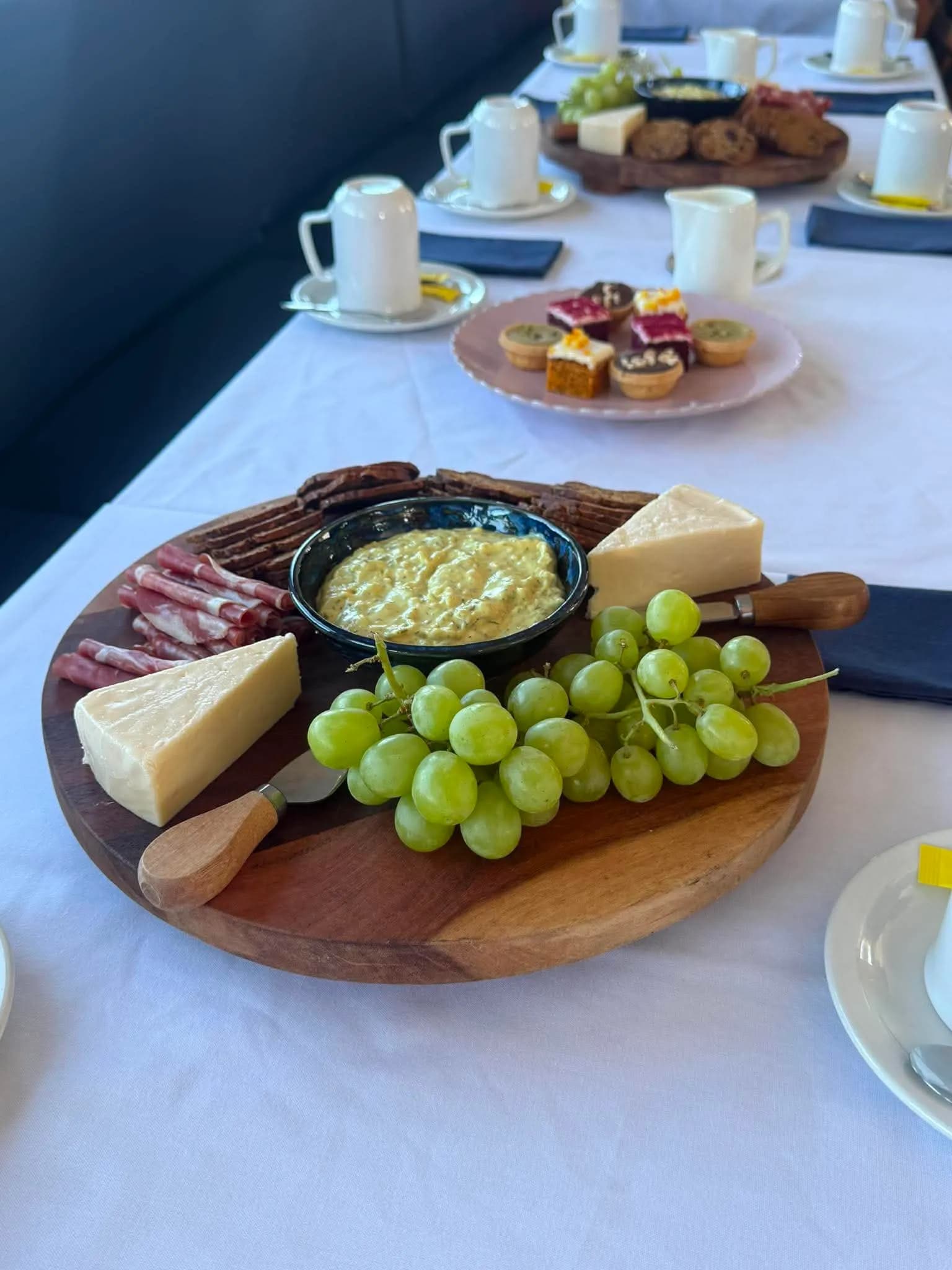 A grazing board of cheese, cured meats, dip and grapes on a white tablecloth aboard MV Mini Munya, with petit fours behind